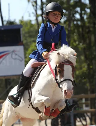 Jeune cavalière portant une veste de concours bleue et une bombe noire, montant un poney blanc et roux à crinière blanche lors d'un saut d'obstacles en compétition
