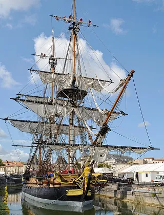 Gros plan sur une grande réplique de frégate à trois mâts et voiles carrées, amarrée dans un port de ville sous un ciel bleu. Des membres d'équipage sont visibles en haut du gréement