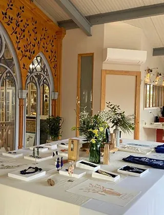 Table de présentation d'objets ou de bijoux, avec des vases de fleurs et des créations étalées sur des supports blancs, dans un espace intérieur lumineux. L'arrière-plan révèle une architecture décorative en bois sculpté avec des fenêtres de style gothique