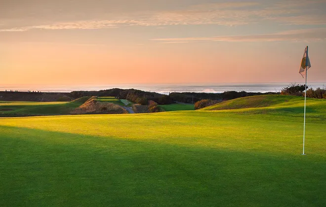 Vue panoramique d'un terrain de golf en bord de mer au coucher du soleil. Le green est éclairé d'une lumière chaude, et un drapeau de golf est visible au premier plan, avec l'océan à l'horizon