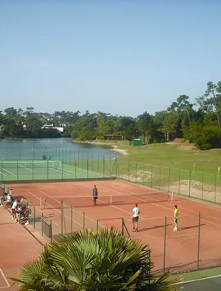 Vue plongeante sur des courts de tennis en terre battue et en gazon synthétique, situés au bord d'un lac. Des personnes jouent au tennis tandis que d'autres sont assises sur les bancs d'observation. L'ensemble est entouré de pins