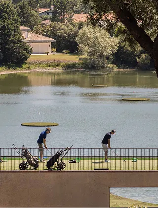 Deux golfeurs s'entraînent sur un practice donnant sur un étang. Ils sont sur une plateforme herbeuse clôturée, avec leurs sacs de golf, et visent des cibles flottantes sur l'eau. Des maisons sont visibles derrière l'étang