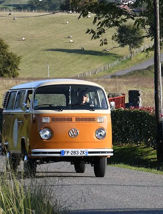 Un van Volkswagen Combi T2 orange et blanc, immatriculé en France, roule sur une petite route de campagne. En arrière-plan, on voit une colline verdoyante avec quelques moutons et une route sinueuse