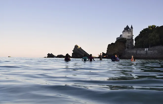 Un groupe de personnes se baignant dans l'océan au coucher du soleil, avec la mer calme au premier plan. À l'arrière-plan, un rocher escarpé surmonté d'une grande villa historique à tourelle, probablement à Biarritz
