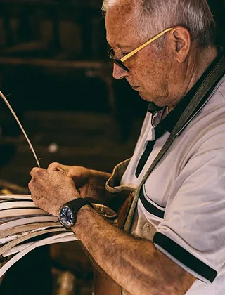 Gros plan sur un artisan plus âgé, portant des lunettes et une montre, concentré sur le tissage ou le tressage de fines lanières de bois ou de fibres