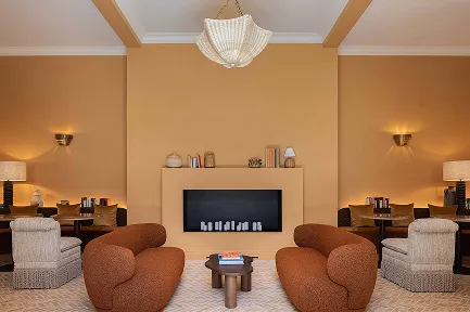 Intérieur d'un salon confortable avec des murs couleur ocre et une cheminée moderne et minimaliste. Deux petits canapés rembourrés en tissu bouclé roux se font face, encadrés par des fauteuils clairs. Une grande suspension en perles est centrée au plafond