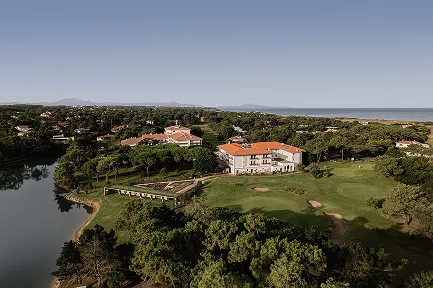 Vue aérienne d'un grand hôtel avec un toit en tuiles rouges, entouré d'un terrain de golf, d'une forêt de pins et d'un plan d'eau. La propriété est située près de la côte, avec la mer et des collines visibles à l'horizon