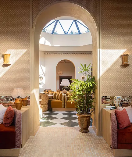 Intérieur d'un hall d'entrée baigné de lumière, décoré avec des matériaux naturels et un style méditerranéen. On y voit un passage sous une arche menant à un salon. Le sol est composé de petits carreaux clairs et d'un damier noir et blanc