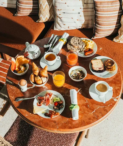 Vue de dessus d'une table ronde en marbre rouge, dressée pour un petit-déjeuner copieux. Elle comporte des œufs brouillés, des croissants, des fruits frais, de la charcuterie et du fromage, des crêpes, des jus de fruits et des boissons chaudes. La table est installée sur une terrasse baignée de soleil, avec des coussins à motifs en arrière-plan