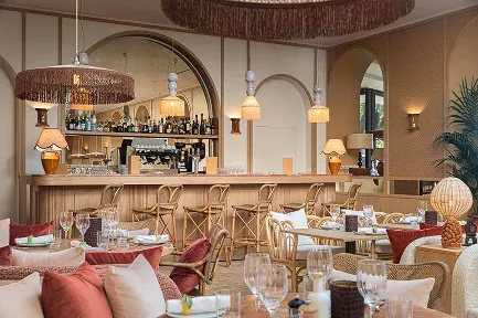 Intérieur d'un bar-restaurant lumineux au style contemporain et bohème. On y voit un grand comptoir en bois clair avec des tabourets hauts en rotin. La salle est décorée avec des suspensions et des luminaires à franges, un grand miroir, des fauteuils en rotin, et des coussins aux tons rose poudré et rouille