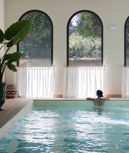 Piscine intérieure avec une femme de dos se reposant au bord de l'eau. Le bassin est éclairé par une série de grandes fenêtres cintrées offrant une vue sur une végétation luxuriante, avec de courts rideaux blancs