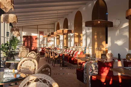 Intérieur d'un grand restaurant lumineux, de style bohème et méditerranéen. Il présente des murs blancs, des arches ocre, un sol en terre cuite et un plafond à poutres apparentes. Le mobilier se compose de chaises et de suspensions en rotin, et de banquettes rouge bordeaux le long des murs.