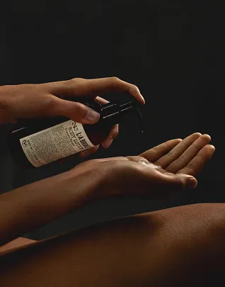 Primer plano de las manos, una de las cuales sostiene un frasco de loción oscura (posiblemente La Bruket Body Lotion) y coloca un poco de producto en la palma de la otra mano, sobre un fondo negro