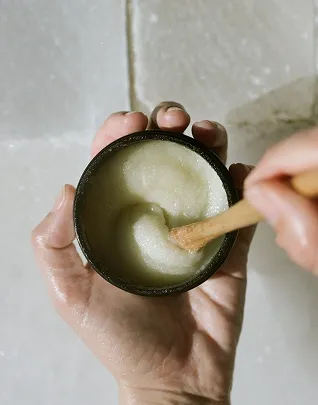 Primer plano de una persona que sostiene un frasco de exfoliante corporal de color claro (posiblemente azúcar o sal) y lo mezcla con una espátula o cuchara pequeña de madera