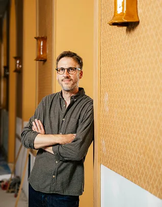hombre de negocios vestido con camisa gris oscuro y gafas, brazos cruzados, apoyado contra una pared discretamente estampada en un pasillo interior.
