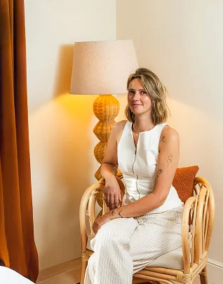 Mujer sonriente con un top blanco sin mangas y pantalones de rayas claras, sentada en un sillón de ratán frente a la pantalla de una lámpara y una cortina de color óxido.