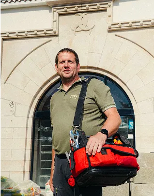hombre de negocios sonriente vestido con un polo y una bolsa de herramientas profesional roja, de pie frente a una fachada de piedra con un arco.