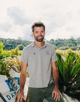 Hombre sonriente vestido con un polo gris y pantalones caqui, de pie entre plantas de vivero y bolsas de fertilizante. El cielo está parcialmente nublado y al fondo se ven árboles y vallas.