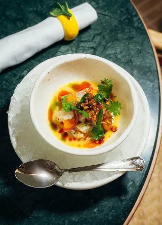 Un petit bol blanc de ceviche, contenant du poisson cru mariné, des tomates, de la coriandre et une sauce jaune orangée, posé sur une plus grande assiette remplie de glace pilée. Une cuillère argentée repose sur la glace à côté du bol, sur une table sombre marbrée de vert