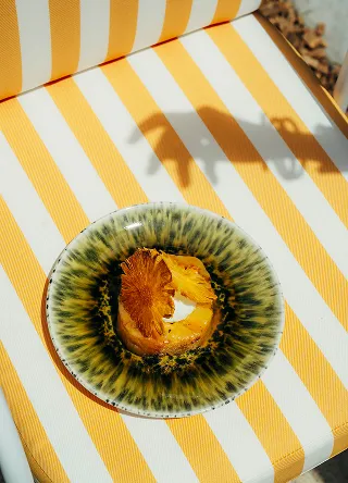 Dessus d'une assiette en céramique à motifs verts et jaunes contenant une tranche d'ananas frais ou grillé, posée sur une chaise longue rayée jaune et blanche, avec l'ombre d'une main visible en haut à droite