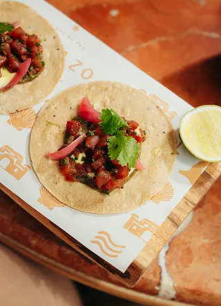 Deux petits tacos de maïs sur une planche de bois et du papier, chacun garni de morceaux de poisson cru mariné (type thon ou ceviche), de fines lanières d'oignon mariné, et d'une feuille de coriandre. Un quartier de lime est visible à droite
