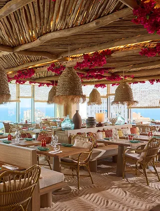 Interior of a seaside or exotic restaurant, with a wooden and straw ceiling, wicker pendants, and red flowers. The wooden tables are set in front of large windows offering views of the sea and the city.