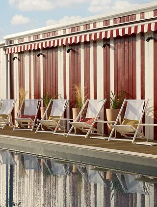 Exterior of a pool with a row of white lounge chairs at the water's edge. The wall of the building behind is decorated with wide vertical red and white stripes, matching the awning above.