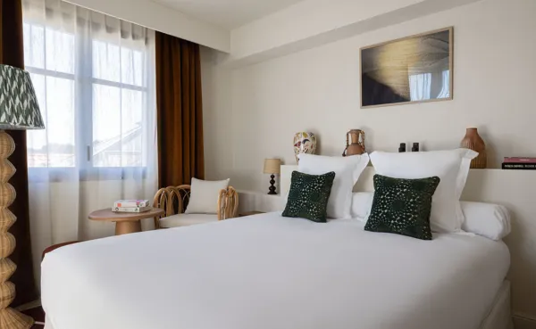Habitación de hotel moderna y luminosa con una cama grande con ropa de cama blanca, cojines decorativos de color verde oscuro y un cabecero con un estante decorado con jarrones. A la izquierda, una mesita auxiliar, un sillón de ratán y una gran ventana con velo enmarcada por cortinas marrones