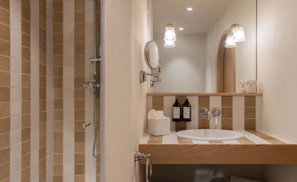 Cuarto de baño moderno de hotel con una ducha con azulejos de rayas verticales de color marrón claro y blanquecino. El mostrador debajo del espejo repite el patrón de rayas con un lavabo incorporado, un espejo de aumento montado en la pared e iluminación decorativa.