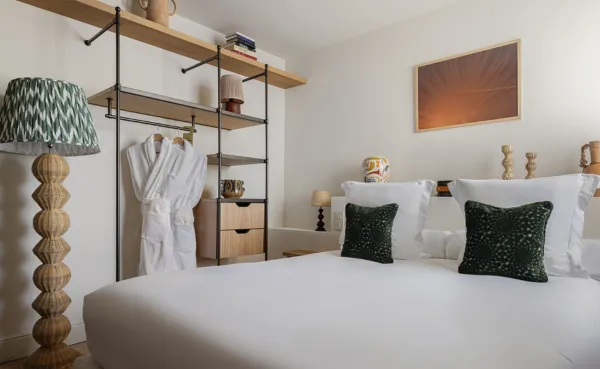 Interior de una luminosa habitación de hotel con una cama grande con ropa de cama blanca, dos cojines decorativos de color verde oscuro y un armario industrial en madera clara y metal del que cuelgan dos albornoces blancos