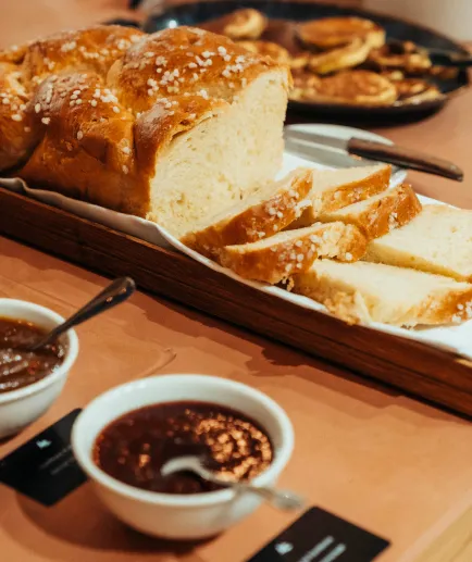 Close-up on a piece of woven and sliced brioche, sprinkled with pearl sugar, presented on a buffet platter. Two bowls of jam or spread are in the foreground.