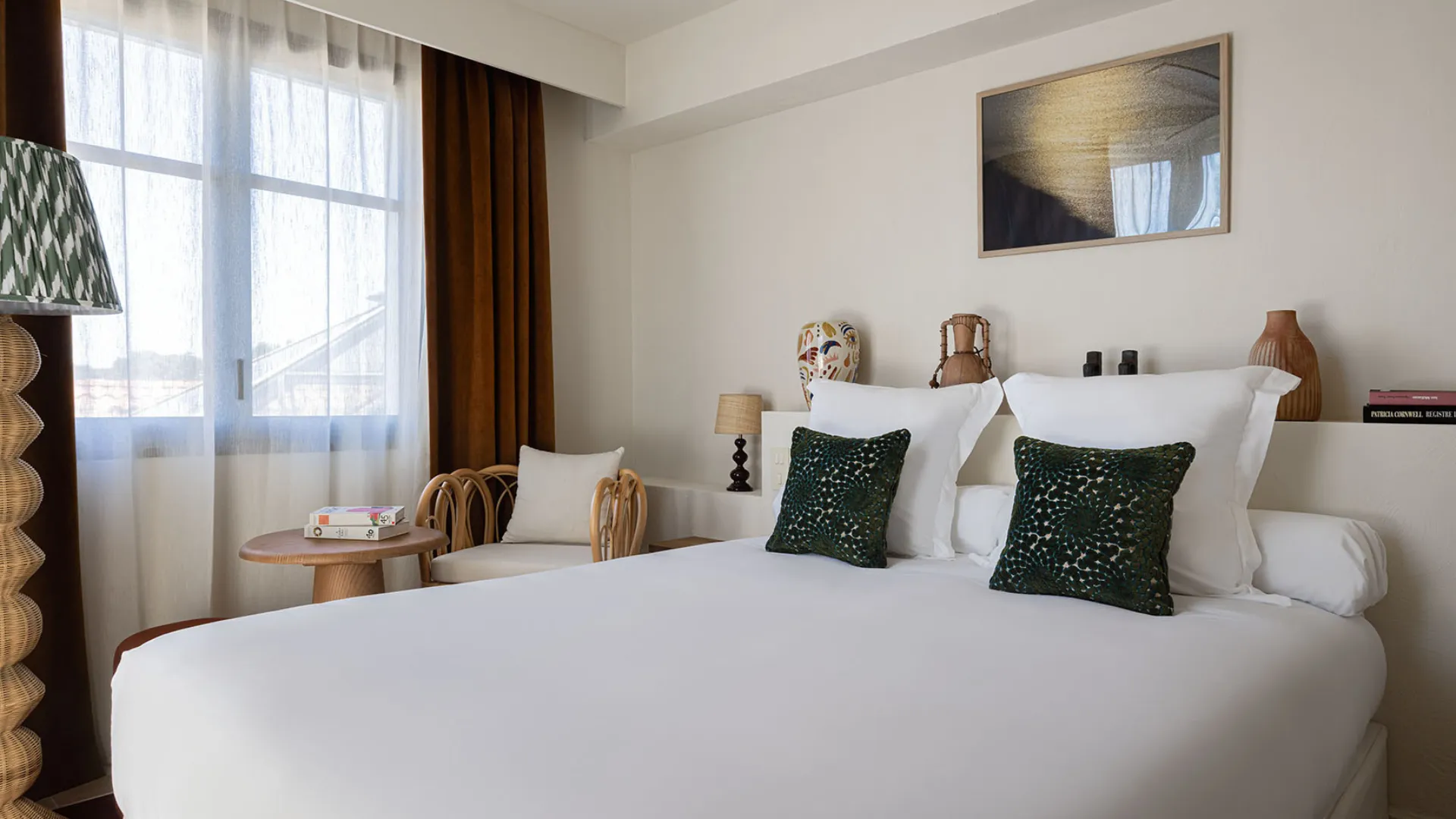 Modern hotel room with a white double bed, wicker floor lamp, industrial shelves, and white bathrobes.