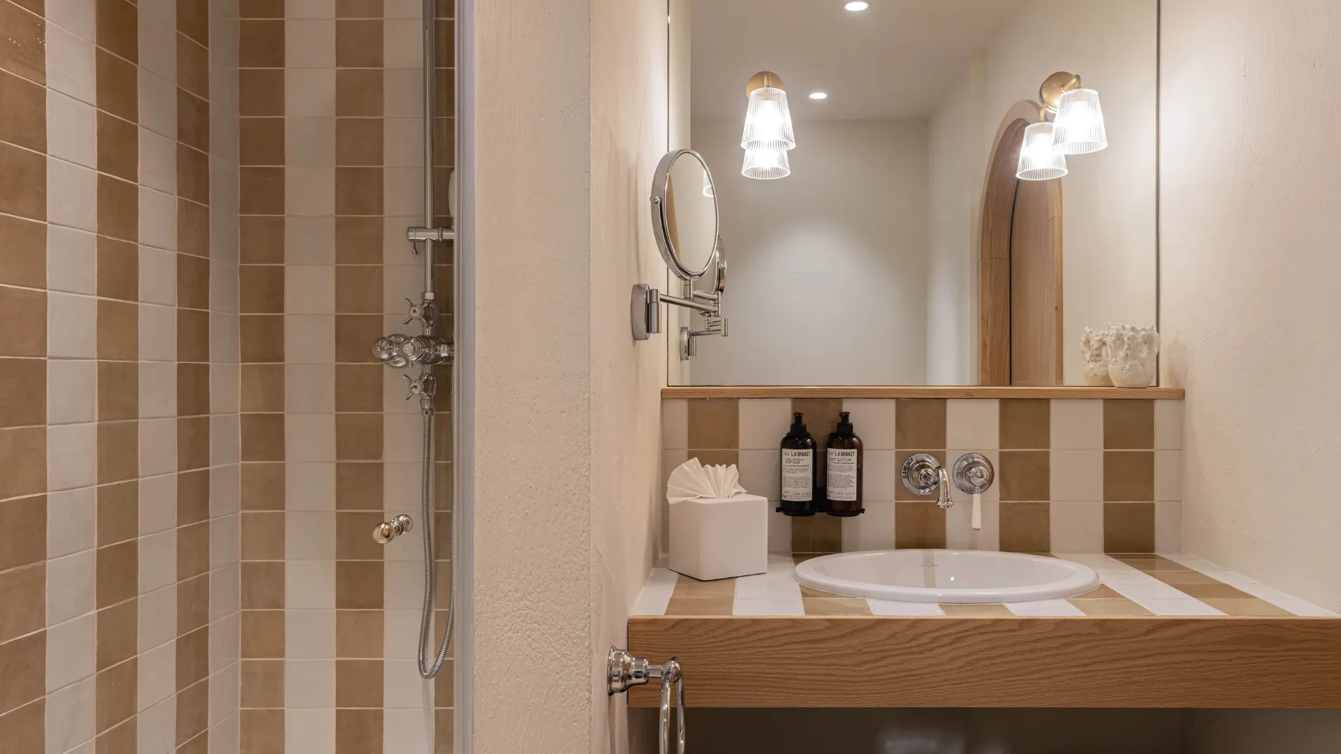 Modern and elegant bathroom with beige and white striped and checkered tiles, a white vessel sink built into a light wood countertop, large mirror, and adjacent shower.