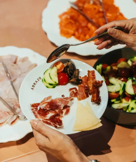 Gros plan sur une assiette blanche avec des morceaux de jambon cru, du bacon croustillant, du fromage et des légumes grillés (concombre, tomates). Une main utilise une cuillère pour ajouter de la nourriture. D'autres plats de charcuterie sont visibles autour de l'assiette