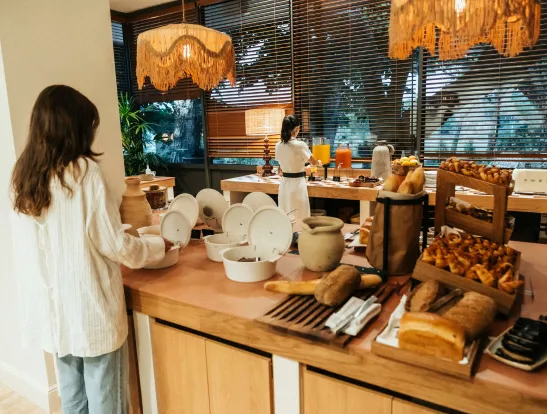 Scène de buffet petit-déjeuner : deux femmes se servent, l'une près des plats chauds couverts et l'autre derrière les distributeurs de jus. Le comptoir est rempli de pains, de viennoiseries, et l'espace est éclairé par des suspensions en paille tressée