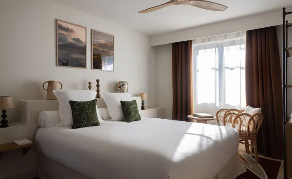 Habitación de hotel elegante y luminosa con una gran cama de lino blanco, cojines decorativos de color verde oscuro y un sillón de ratán junto a la ventana. Se ve un ventilador de techo y el espacio está decorado con cortinas marrones y dos obras de arte enmarcadas sobre la cama.