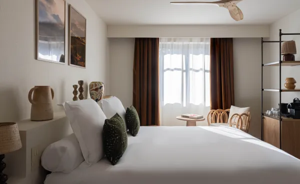 Habitación de hotel moderna y luminosa con una cama grande de lino blanco, cojines decorativos de color verde oscuro y un ventilador de techo. La cabecera está decorada con jarrones altos y el espacio junto a la ventana está amueblado con una silla de ratán