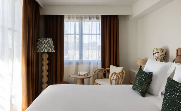 Habitación de hotel moderna y luminosa con una gran cama de lino blanco, cojines de color verde oscuro y una gran lámpara de pie decorativa con una pantalla estampada y una base de paja tejida. La ventana está rodeada de cortinas marrones