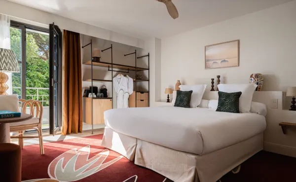 Amplia habitación de hotel con una cama grande de lino blanco, un ventilador de techo y un armario abierto de estilo industrial con estantes y un albornoz colgante. La puerta del patio conduce a un balcón rodeado de vegetación, y hay un sillón de ratán cerca de la mesita de noche