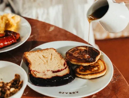 Close-up of a person pouring maple syrup from a small white pitcher onto two pancakes and a slice of fruit bread, served on a white plate.