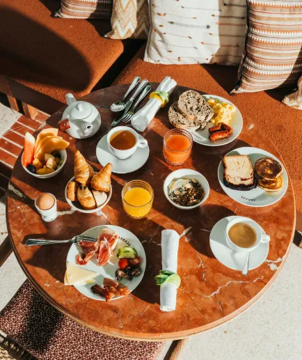 Top view of a large breakfast for two, served on a round red marble table. The display includes eggs, fruit, coffee, coffee, juice, juice, pastries, pancakes, charcuterie, and toast.