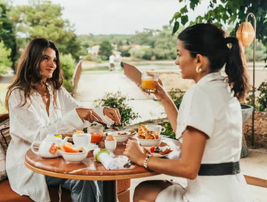 Two friends have breakfast on the terrace, sitting at a round table with eggs, fruit and pastries.