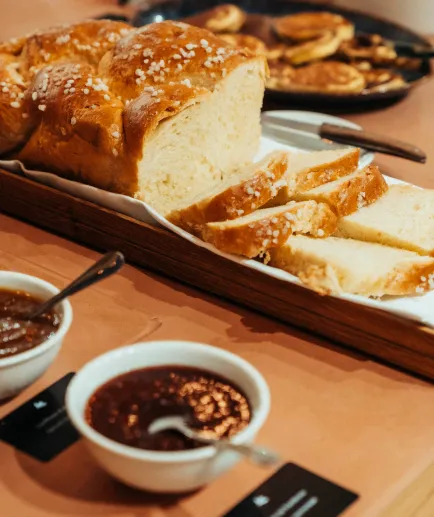 Close-up on a brioche braid cut into slices and sprinkled with pearl sugar, presented on a wooden tray. Two bowls of jam or spread are visible in the foreground.