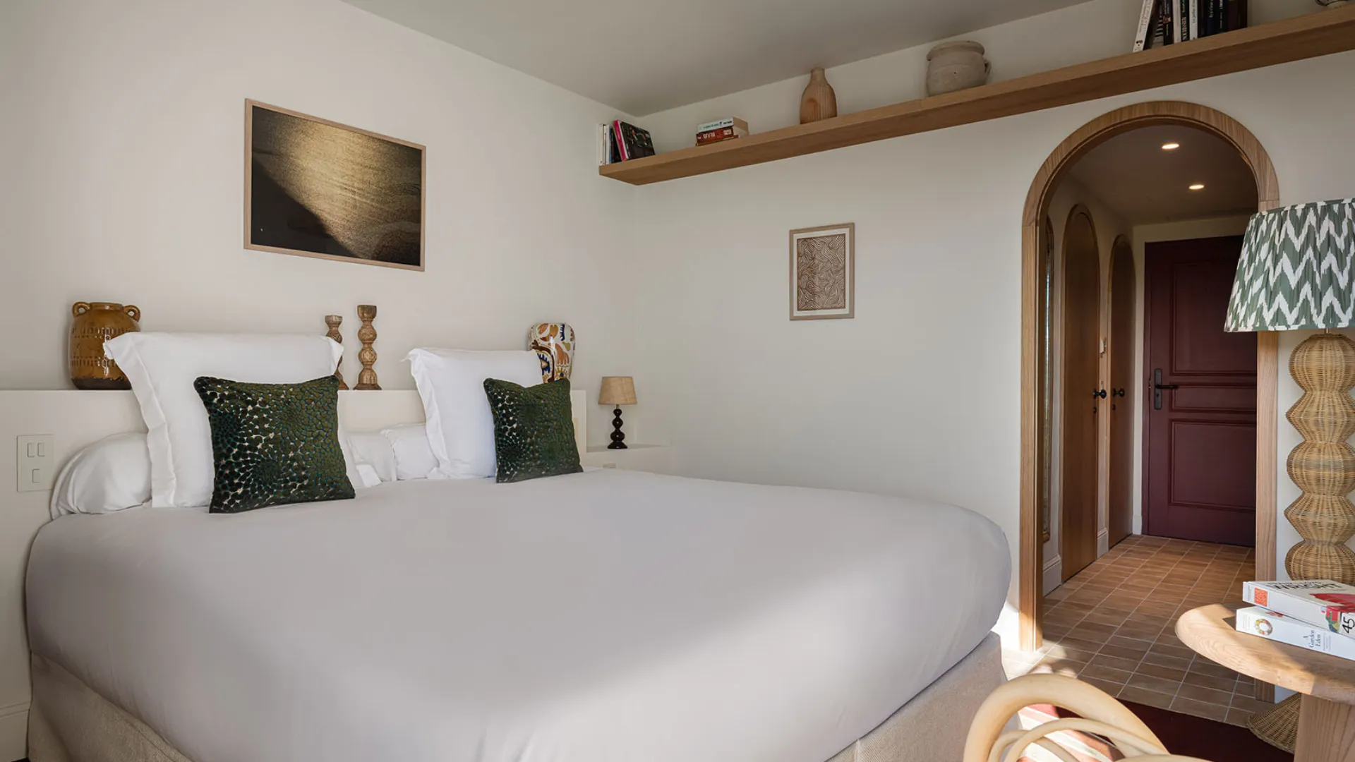 Grande chambre d'hôtel au décor moderne, lit double blanc et entrée arquée vers la salle de bain ou le dressing.