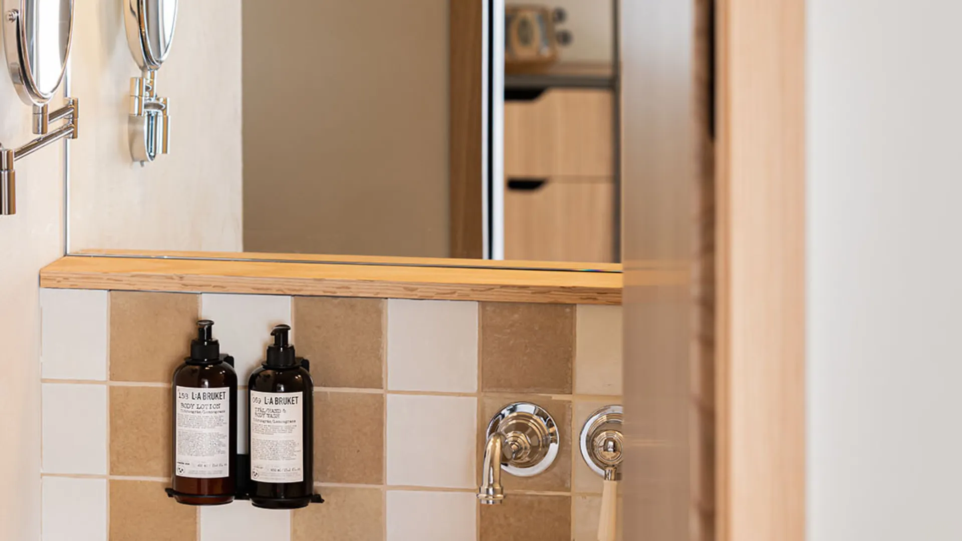 Detail of a modern bathroom featuring beige and white square tiles, a wooden shelf edge, and two L:A BRUKET toiletries.