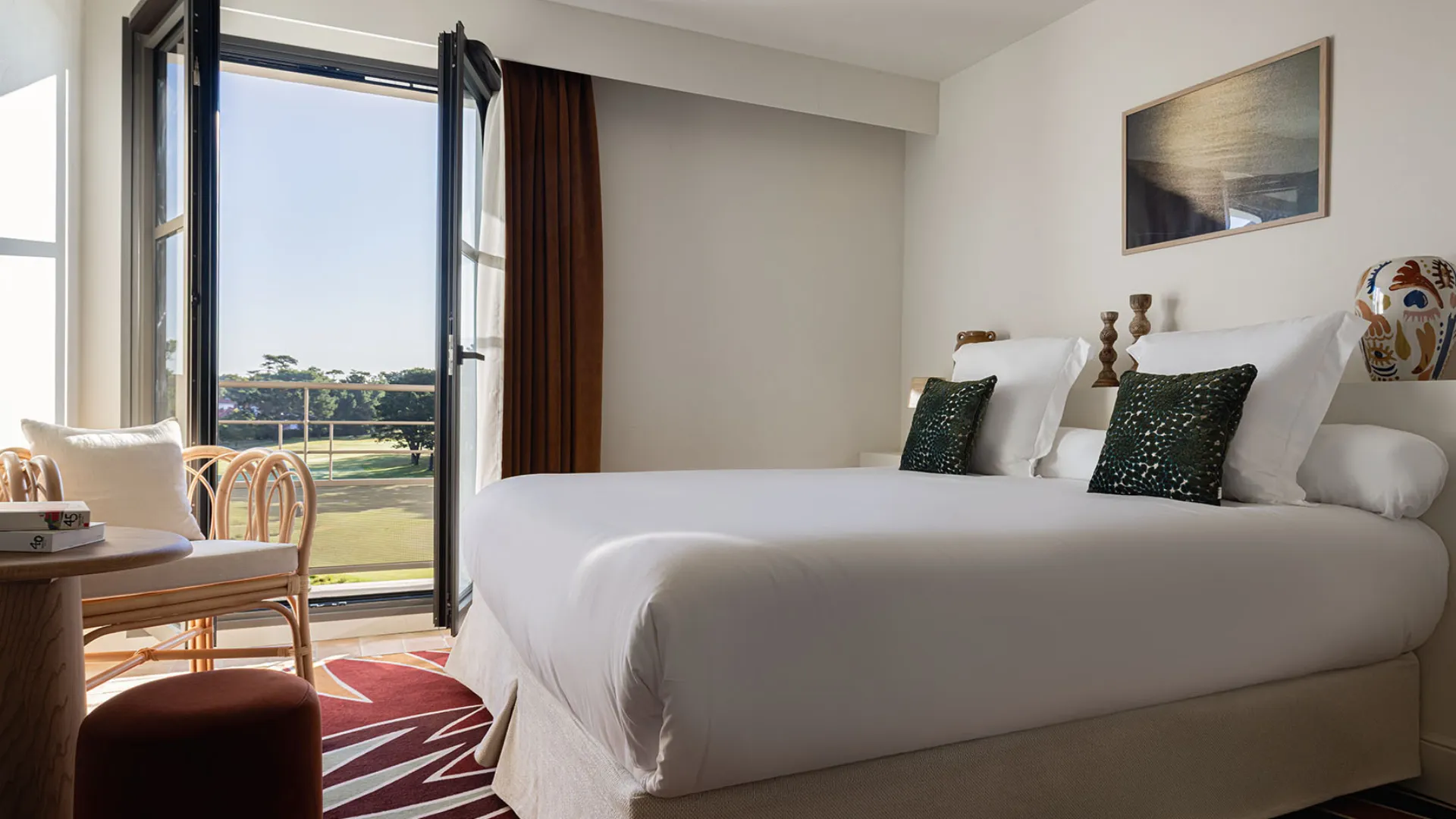 Elegant hotel room with white bedding, brown curtains, a rattan armchair, and access to a balcony overlooking a golf course.