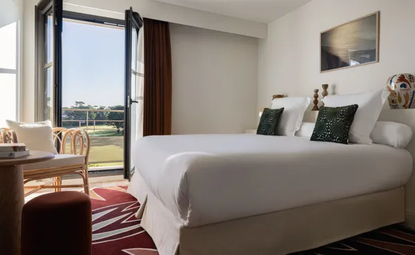 Habitación de hotel luminosa con una cama doble grande y una ventana francesa con vistas a un balcón y un césped verde. Junto a la ventana hay un sillón de ratán y una mesa redonda pequeña. La habitación está decorada con cojines de color verde oscuro y una alfombra roja estampada.
