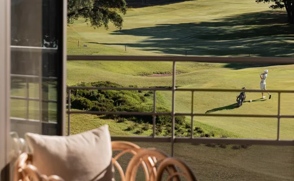 Vista de un campo de golf ondulado desde el balcón de una habitación de hotel. Se ve a un golfista pateando una pelota y su bolsa de golf. Los muebles del balcón, que incluyen un sillón y un cojín de ratán, se ven en primer plano.