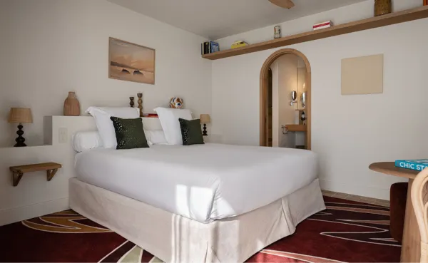 Habitación de hotel moderna y luminosa con una cama grande con ropa de cama blanca, cojines de color verde oscuro y un cabecero con mesitas de noche. Un arco decorativo conduce al baño y un largo estante de madera cuelga sobre la cama