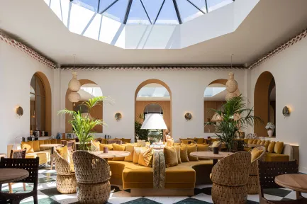Intérieur d'un salon spacieux sous une grande verrière, baigné de lumière naturelle. L'espace présente un décor méditerranéen et chaleureux, avec un grand canapé d'angle couleur moutarde, des fauteuils en paille tressée et des tables basses. Le sol est un damier carrelé vert et blanc, et des arches ocre encadrent l'arrière-plan